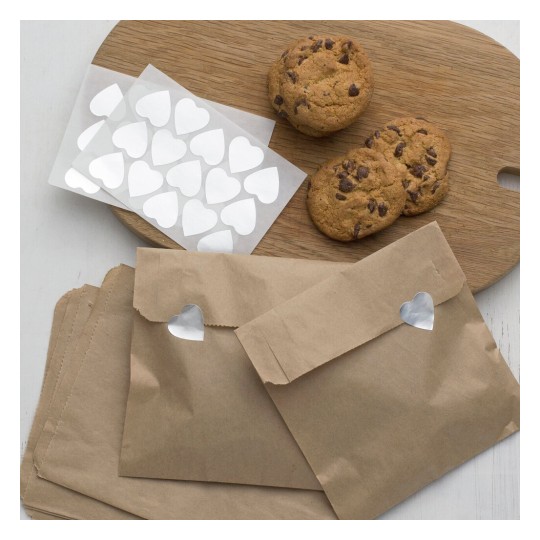 The image features chocolate chip cookies placed on a round wooden cutting board, accompanied by sheets of heart-shaped stickers. In the foreground, there are brown envelopes decorated with heart-shaped cutouts.