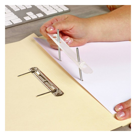 An individual's hand is holding a clear plastic tool with screws, placing it through a sheet of paper inside a folder. A metal binder is visible in the foreground, with a keyboard in the background.
