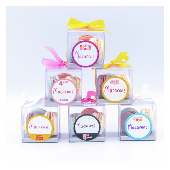 The image features a collection of six clear boxes containing colorful macarons, each with decorative labels and ribbons. The labels include phrases like 'homemade', 'for you', and 'just 4 you'. The macarons are various pastel colors, adding to the appealing presentation.