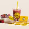 On a neutral background there is takeaway plastic smoothie cup, a plastic bowl container filled with yoghurt and fruit. A pile of business cards, and a sheet of labels with the cafes branding Love Eat