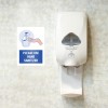 Soap dispenser on a bathroom wall with a sign stuck next to it saying Please use hand sanitizer.