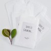 A group of small white paper bags, likely seed packets, with glittery labels on them. The label on the front bag reads "LOVE IS GROWING..." with the names "Henry + Susanna" and "October 11" below. A small sprig of green leaves is visible on the left side of the image.