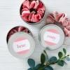 Three round, silver tins with velvet scrunchies inside. The top of the tin on the left has a label with a pink diagonal stripe and the word "luxe" written in pink. The tin on the right has an identical label with the word "cozy" written in pink. One of the scrunchies is spilling out of a tin. A sprig of green leaves is visible in the foreground.