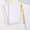 The image shows a collection of blank sheets of paper arranged on a desk, including various colored sheets such as yellow and orange. There is a yellow pencil and a keyboard in the background, along with a calculator and a sticky note.
