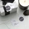 The image features a beautifully wrapped gift with a black ribbon, accompanied by several gift tags. The tags display messages like 'smile' and 'just because I care' along with decorative elements. A lit candle and smooth stones are also visible in the composition, enhancing a serene atmosphere.