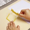 The image shows a person's hand using a yellow tool or clip to hold a piece of white paper against a tan background, likely for some kind of organizational or crafting purpose. A keyboard is visible in the background, suggesting this is a workspace.