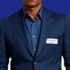 The image shows a man in a dark blue blazer and shirt, wearing a name tag from Universal. The background is a solid color.
