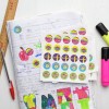 The image features a colorful collection of reward stickers laid on top of school notebooks filled with handwritten notes and drawings. The notebooks display various levels of praise, such as 'Very Good,' 'Excellent,' and 'Fantastic.' A ruler and some pens are also present, indicating a student's workspace.