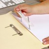 An individual's hand is holding a clear plastic tool with screws, placing it through a sheet of paper inside a folder. A metal binder is visible in the foreground, with a keyboard in the background.