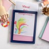 The image features a colorful visual arts assignment folder with vibrant splashes of paint on the cover. Surrounding items include a wooden mannequin, a necklace with colorful beads, colored pencils, paint containers, and stacked books, all arranged on a light-colored surface.