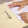 The image shows a person’s hands working with a binder clip on a stack of papers. One hand is securing paper with a clasp, while next to it, there's a metal binder clip positioned on a light-colored folder.